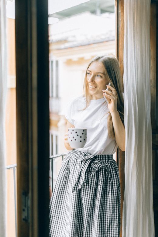 A smiling woman on a phone call by a bright window, holding a mug